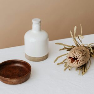 Elegant composition featuring a white bottle, wooden bowl, and dried flower on a table.