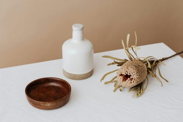 Elegant composition featuring a white bottle, wooden bowl, and dried flower on a table.